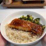 A white bowl filled with steamed rice, glazed salmon fillet with sauce, and roasted broccoli, held in someones hands on a white countertop.