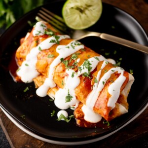 A plate with two enchiladas topped with red sauce, white crema, and chopped cilantro, with a fork and a halved lime in the background.