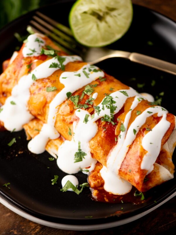 A plate with two enchiladas topped with red sauce, white crema, and chopped cilantro, with a fork and a halved lime in the background.