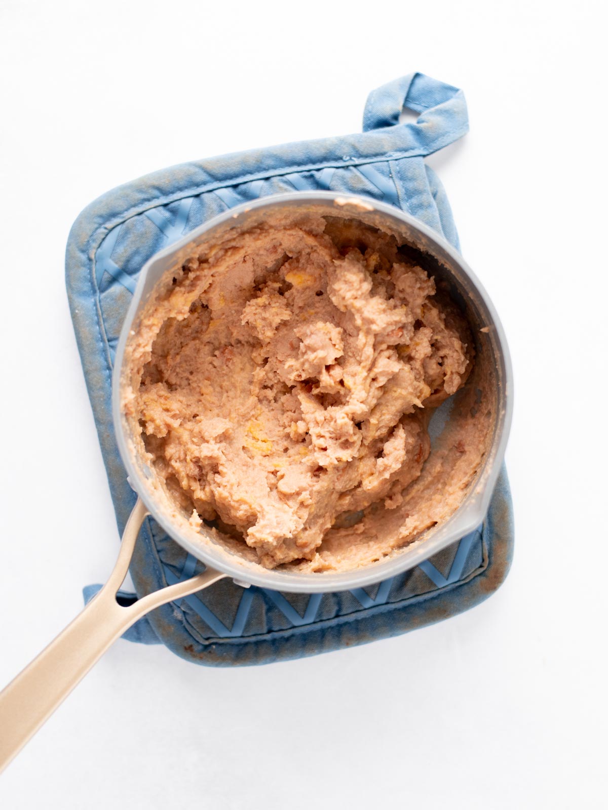 A saucepan filled with creamy refried beans sits on a blue quilted oven mitt, all placed on a white background.