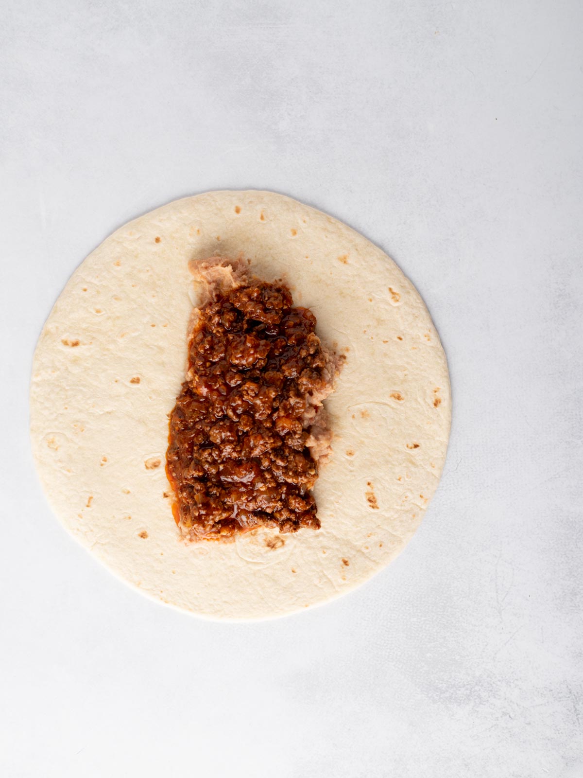 A flour tortilla on a light surface topped with a layer of refried beans and seasoned ground beef filling in the center, ready to be wrapped.