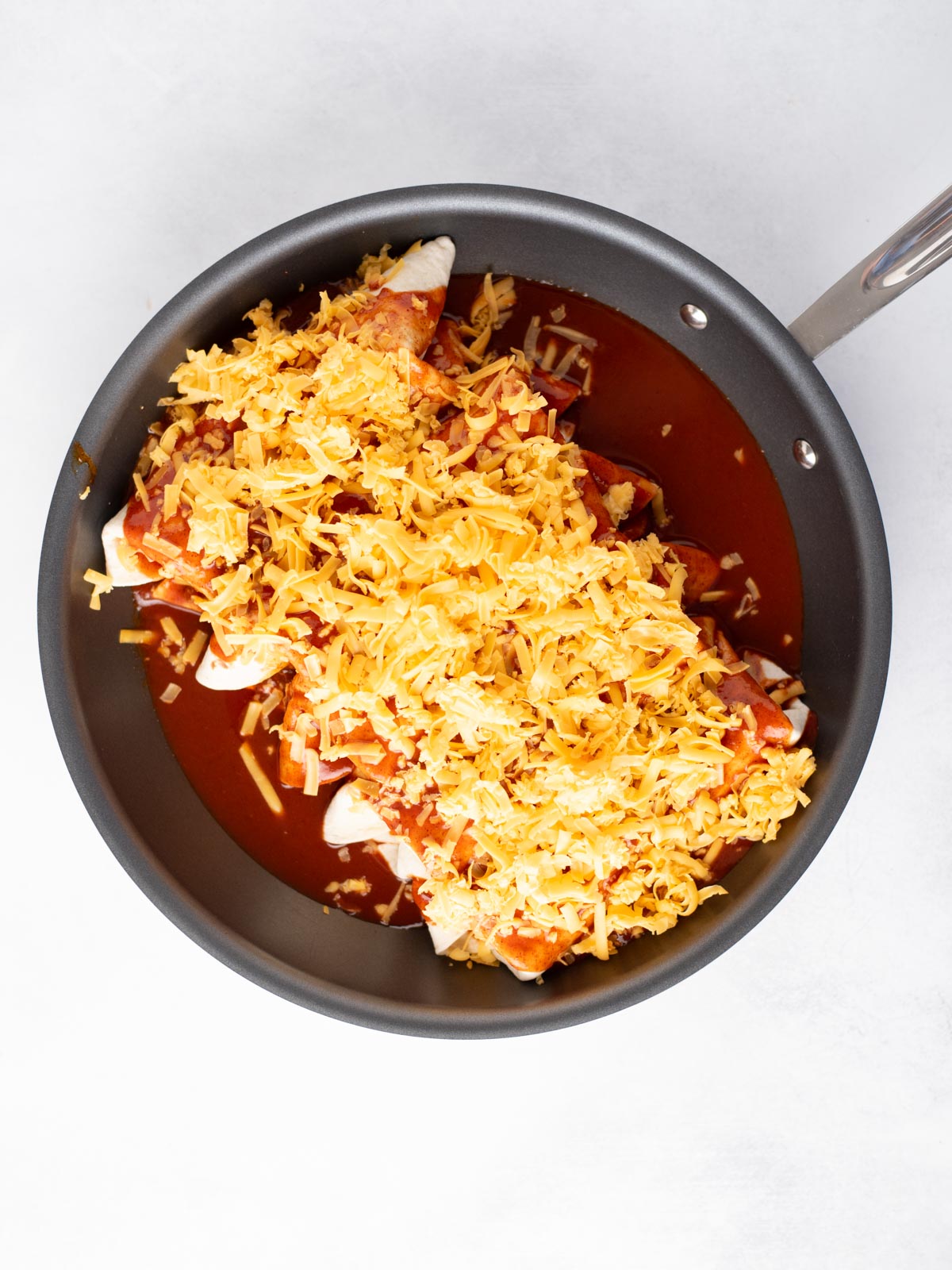 A nonstick skillet filled with uncooked enchiladas, covered in red sauce and topped with shredded cheddar cheese, on a white background.