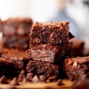 A close-up of a stack of rich, fudgy brownies with a crackly top, surrounded by chocolate chips on a wooden surface. More brownies are blurred in the background.