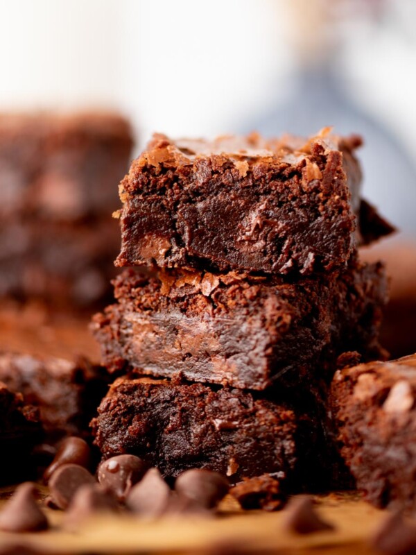 A close-up of a stack of rich, fudgy brownies with a crackly top, surrounded by chocolate chips on a wooden surface. More brownies are blurred in the background.