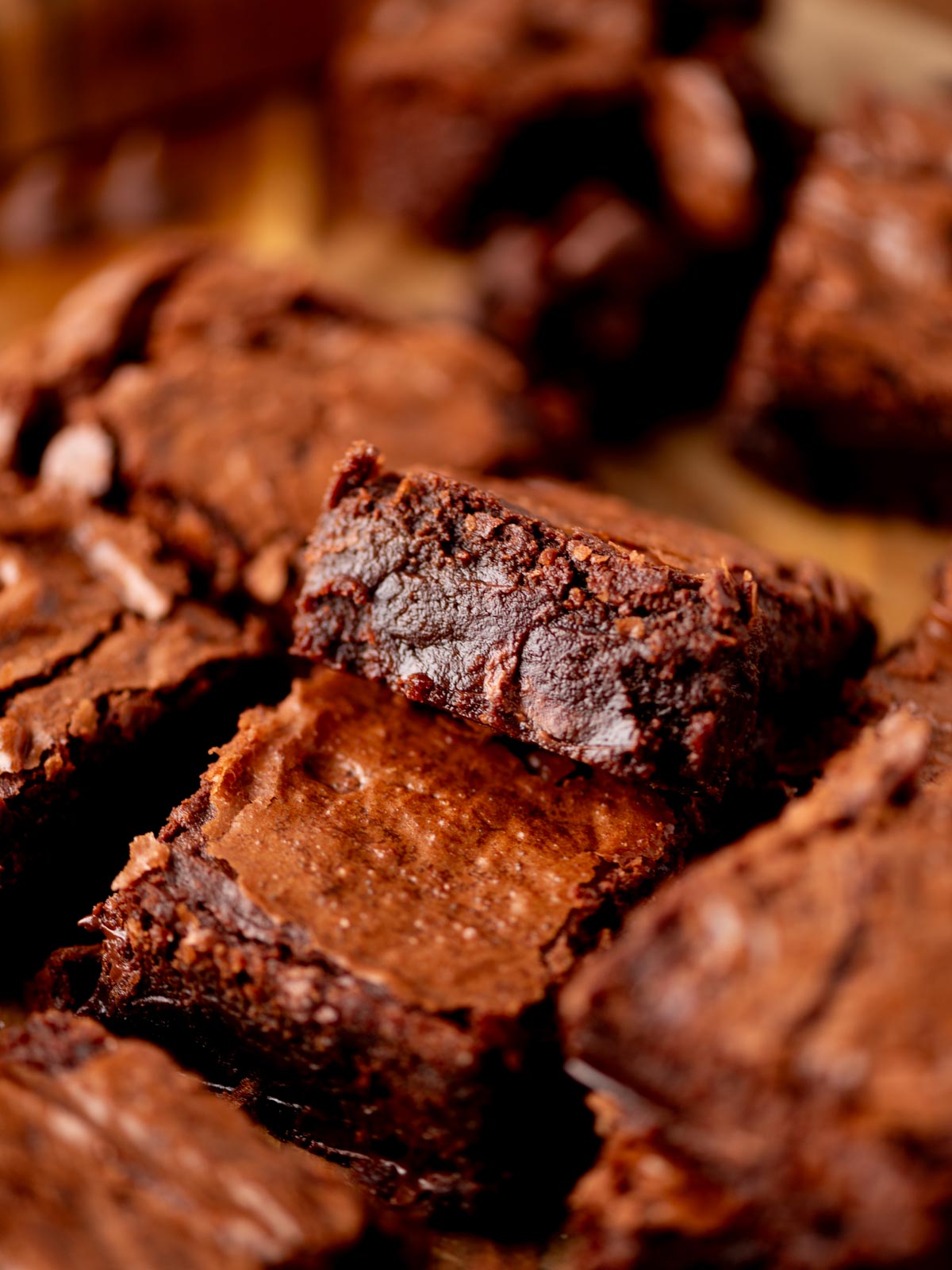 Close-up of rich, fudgy chocolate brownies with a crackly top, stacked and surrounded by other brownie pieces, showcasing their moist, dense texture.