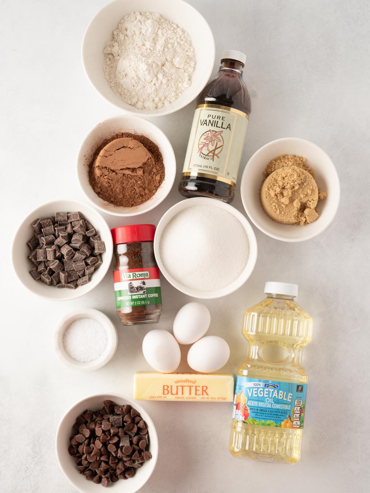 Assorted baking ingredients on a white surface: flour, vanilla extract, brown and white sugar, cocoa powder, instant coffee, chocolate chunks, chocolate chips, eggs, butter, vegetable oil, and salt.