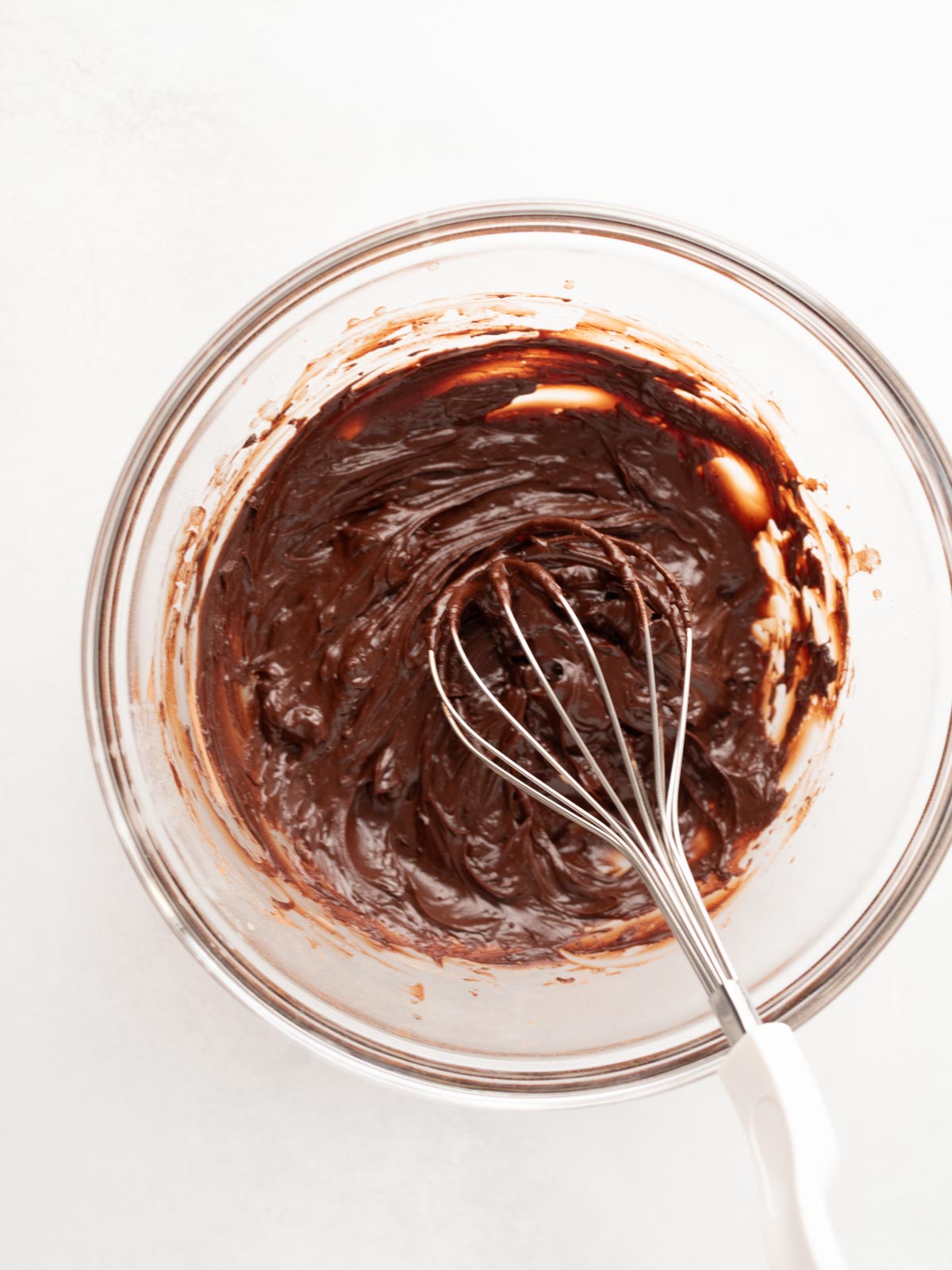 A glass bowl filled with thick chocolate batter being mixed with a metal whisk on a white surface.