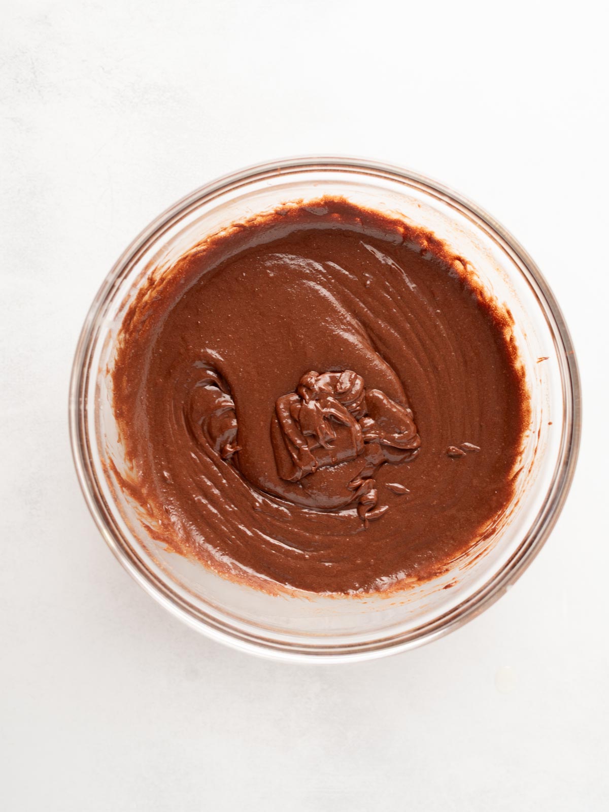 A glass bowl filled with smooth, rich chocolate batter sits on a white surface. The batter has been stirred, leaving visible swirls and a creamy texture.