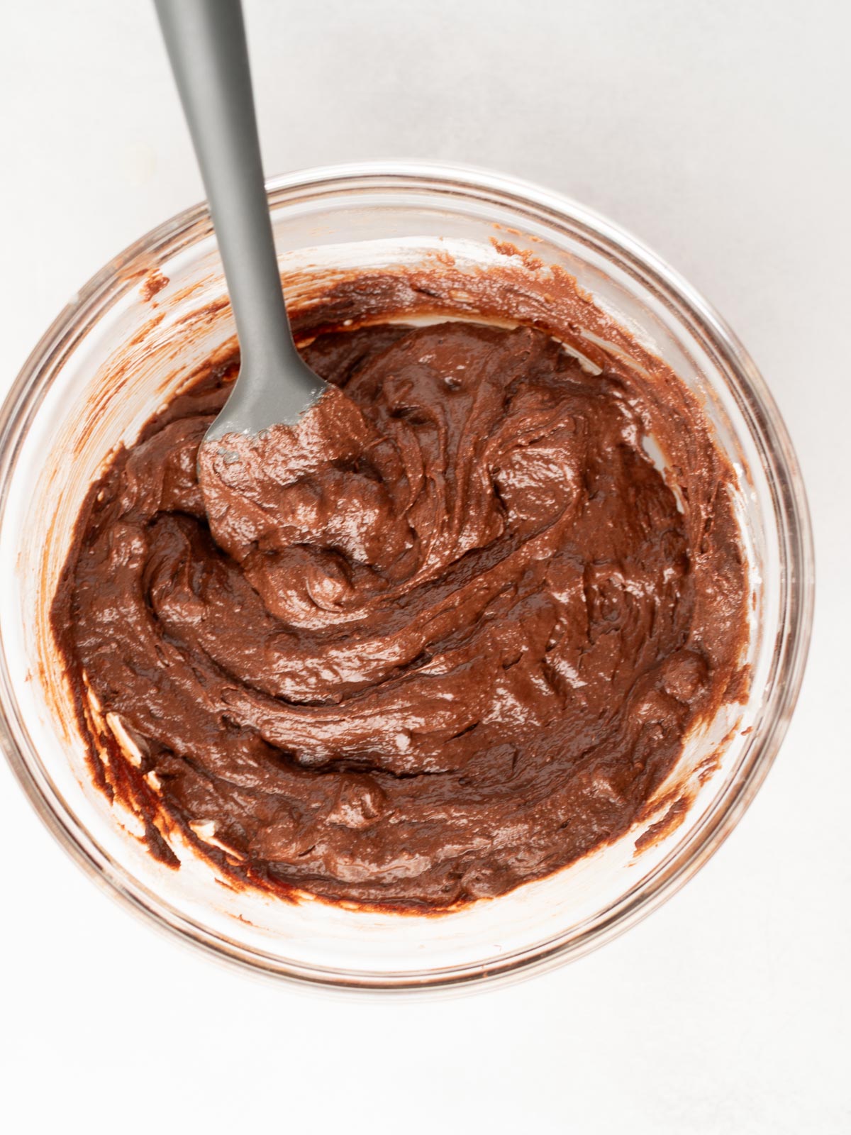 A glass bowl filled with thick, swirled chocolate brownie batter. A spoon rests inside the bowl, partially covered in the rich, dark batter, on a light-colored surface.