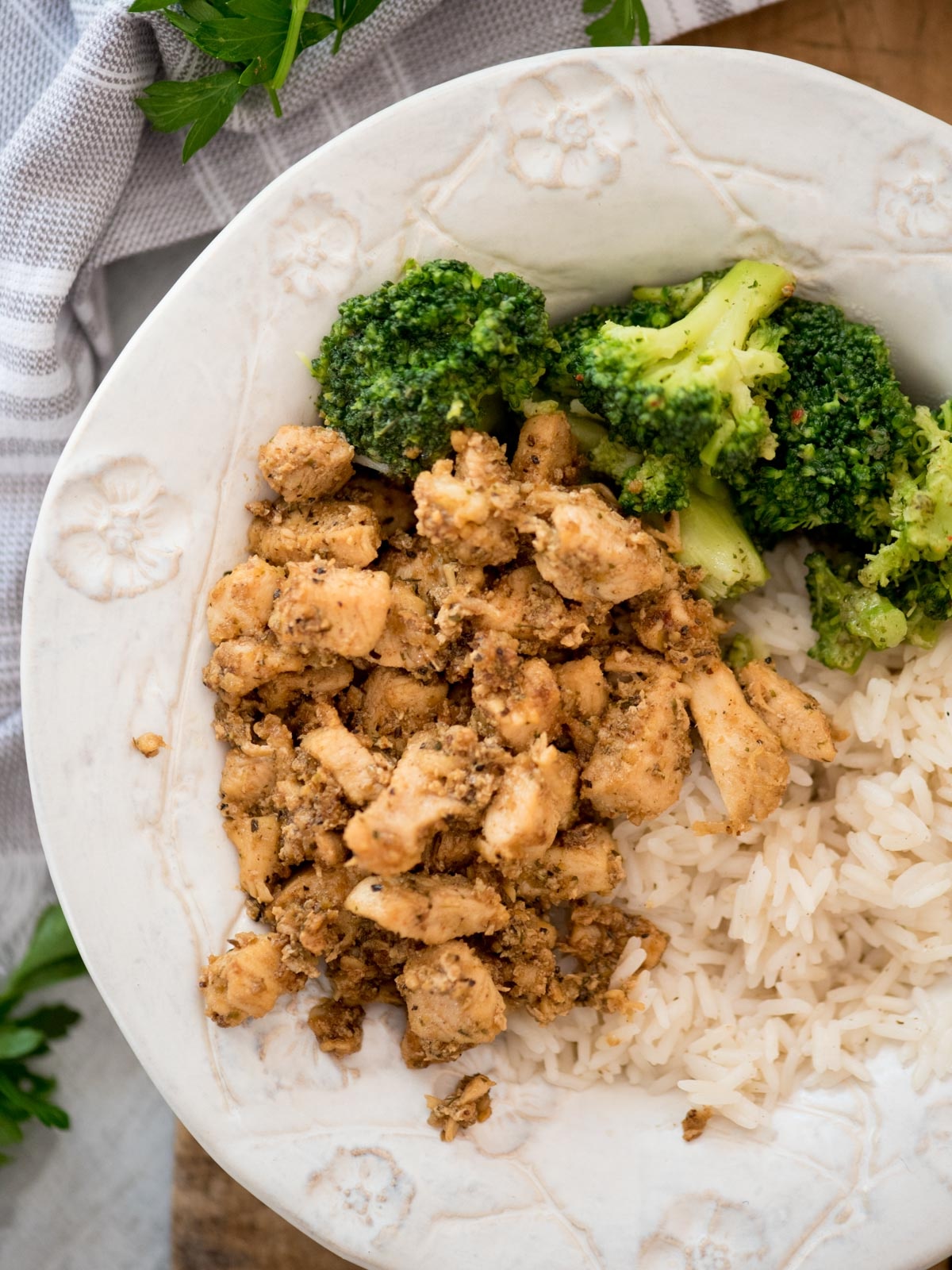 A plate with seasoned diced chicken, steamed broccoli, and white rice, served on a patterned white dish with a cloth napkin and parsley nearby.