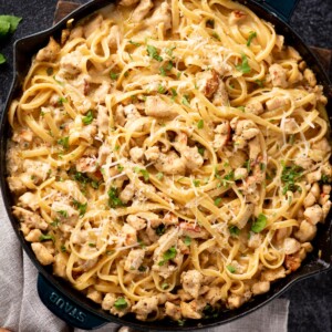 A skillet filled with creamy chicken pasta, topped with chopped herbs and grated cheese, featuring linguine noodles and pieces of cooked chicken.