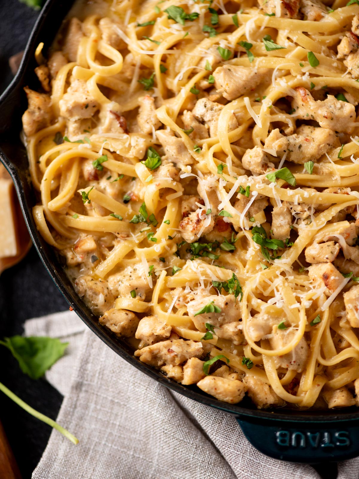 A close-up of creamy chicken fettuccine pasta in a black pan, topped with chopped parsley and shredded parmesan cheese. A beige napkin and some green herbs are partially visible beside the pan.