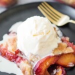 A serving of peach cobbler topped with vanilla ice cream sits on a black plate, with fresh peach slices visible and a fork beside it. A text overlay reads easy fresh peach cobbler sweetlysplendid.com.