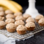 Mini banana muffins are arranged on a cooling rack, with several ripe bananas and a white container blurred in the background. The scene is set on a dark surface with a light-colored cloth underneath the rack.