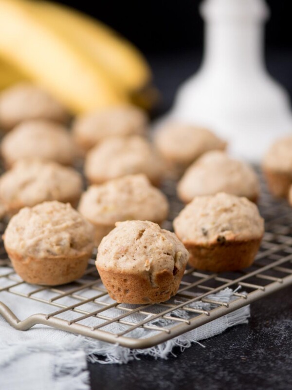 Mini banana muffins are arranged on a cooling rack, with several ripe bananas and a white container blurred in the background. The scene is set on a dark surface with a light-colored cloth underneath the rack.