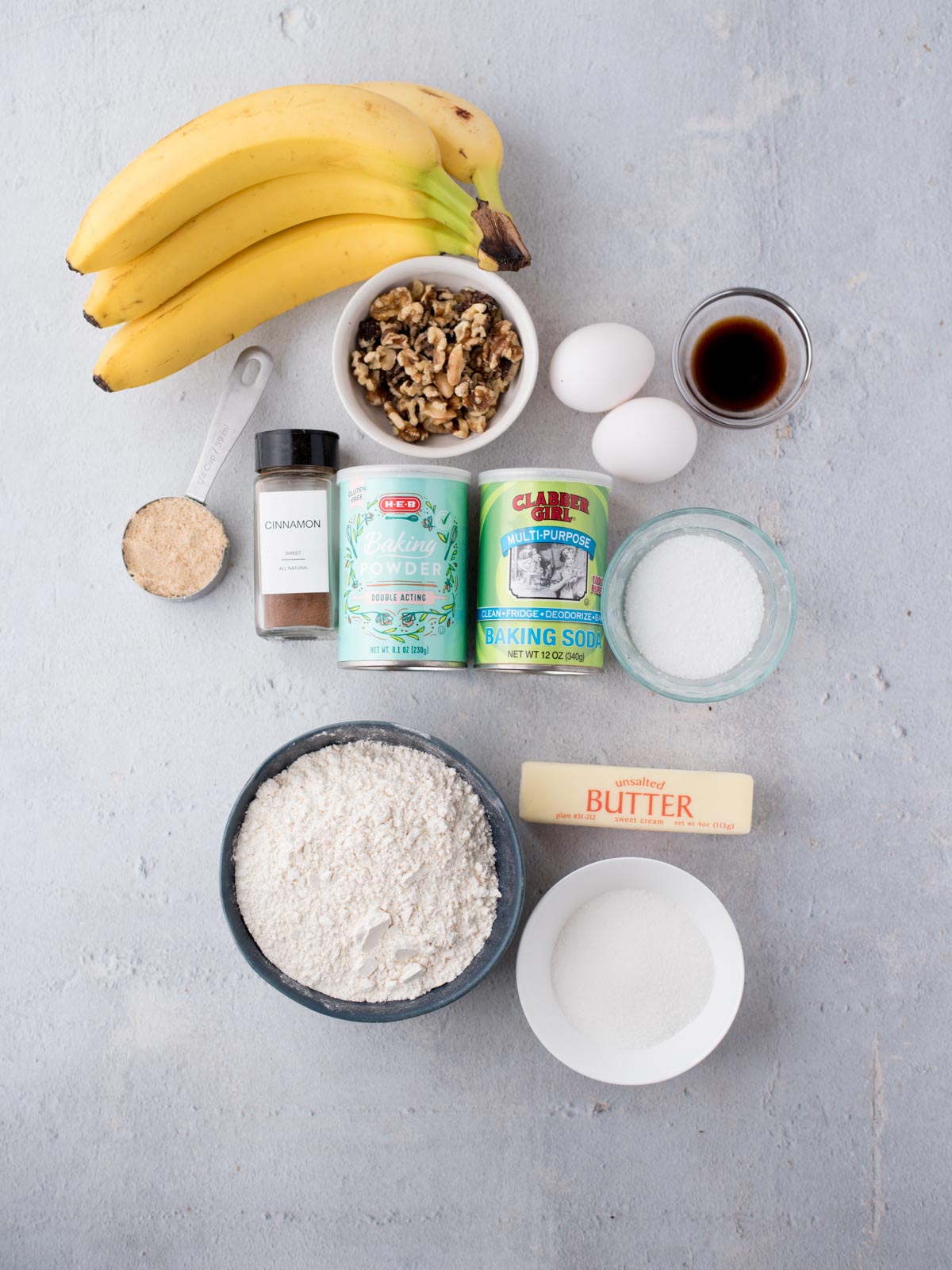 Top-down view of ingredients for banana bread: three bananas, chopped walnuts, two eggs, vanilla extract, baking powder, baking soda, brown sugar, cinnamon, white flour, granulated sugar, and a stick of butter.