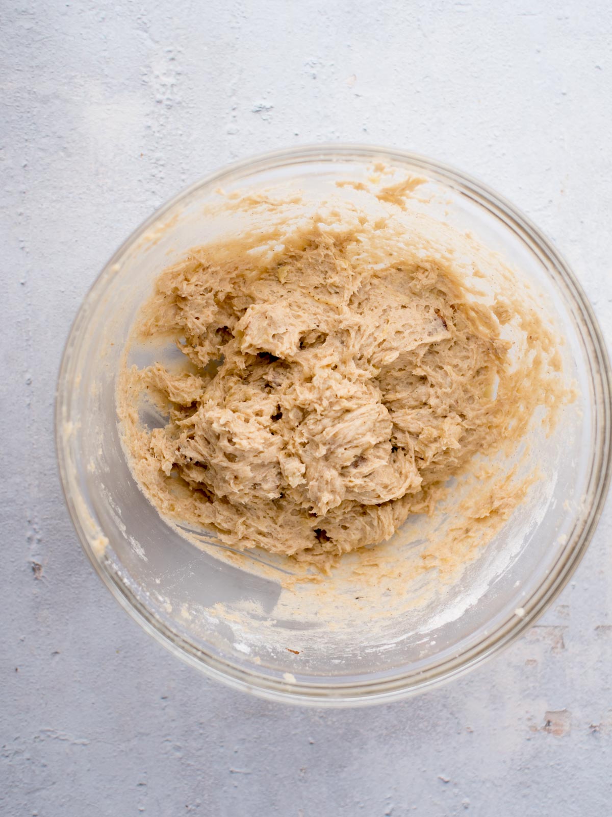 A glass bowl containing thick, creamy cookie dough sits on a light-colored, textured surface. The dough appears partially mixed, with a slightly lumpy texture.