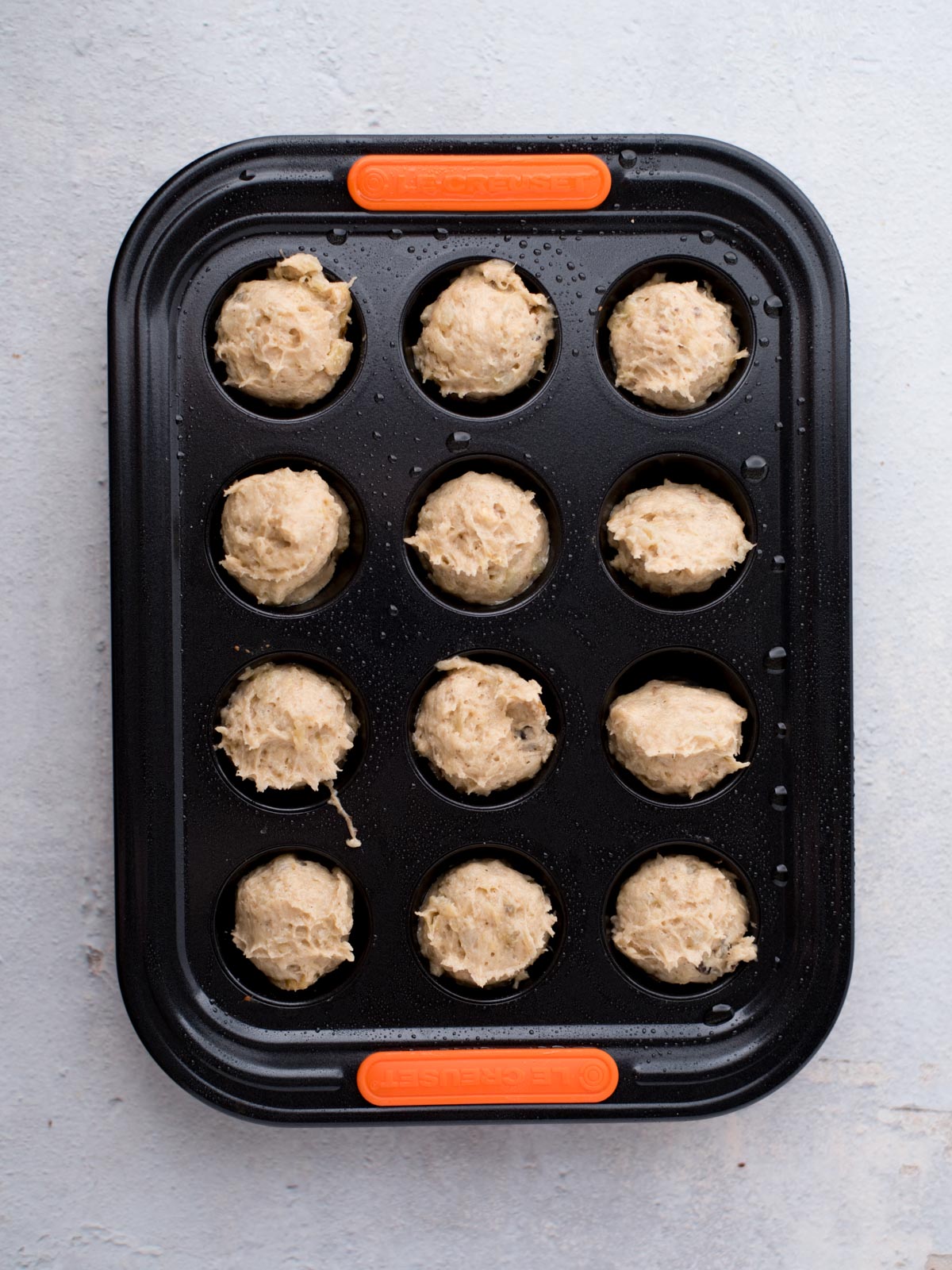 A black muffin tin with twelve round dough balls placed in each cup, ready for baking, sits on a light gray surface. The tin has orange silicone grips on the short ends.