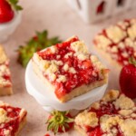 A square strawberry crumble bar sits on a small white pedestal, topped with bright red strawberry filling and golden crumb topping. Other bars and fresh strawberries are scattered around on a light surface.