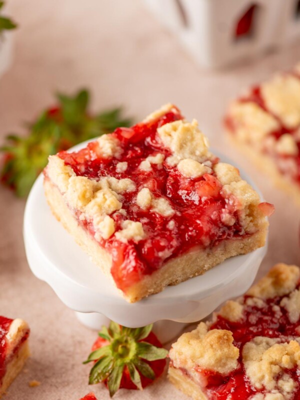 A square strawberry crumble bar sits on a small white pedestal, topped with bright red strawberry filling and golden crumb topping. Other bars and fresh strawberries are scattered around on a light surface.