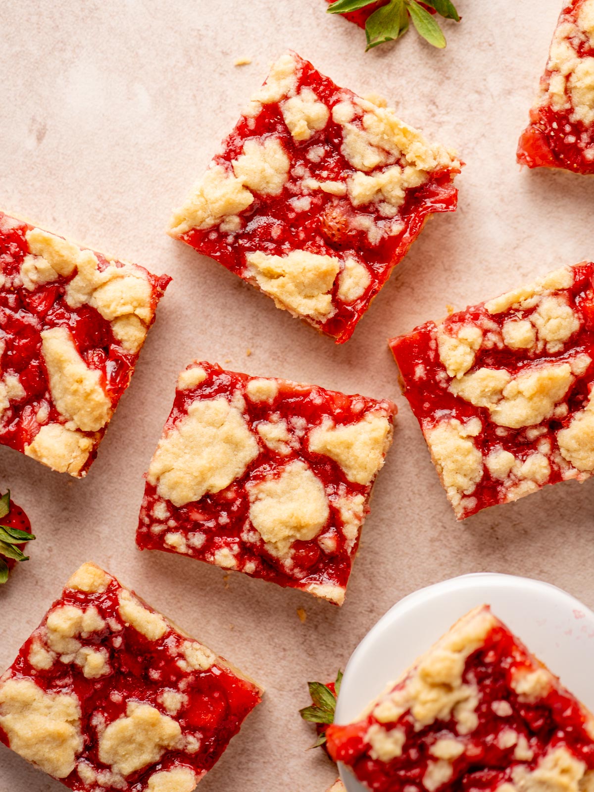 Top-down view of several square strawberry crumble bars with a golden crumb topping on a light surface, surrounded by a few fresh strawberry leaves.