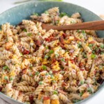 A bowl of creamy rotini pasta mixed with peas, bacon bits, shredded cheese, and garnished with chopped green onions, with a wooden spoon resting in the bowl.