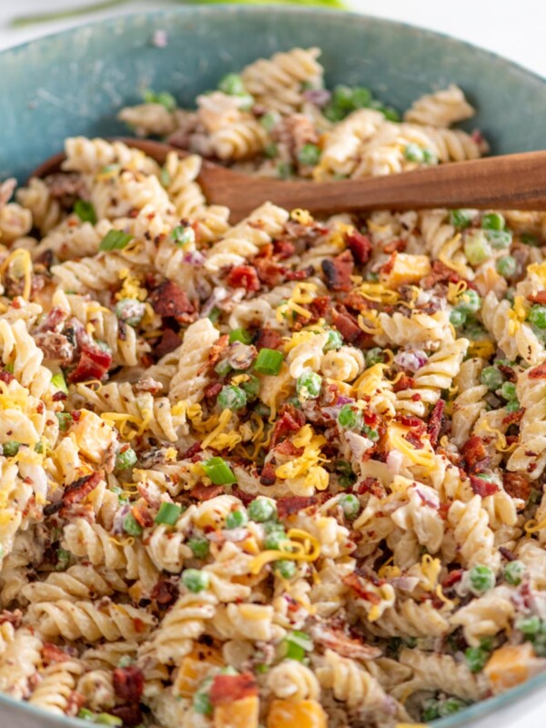 A bowl of creamy rotini pasta mixed with peas, bacon bits, shredded cheese, and garnished with chopped green onions, with a wooden spoon resting in the bowl.
