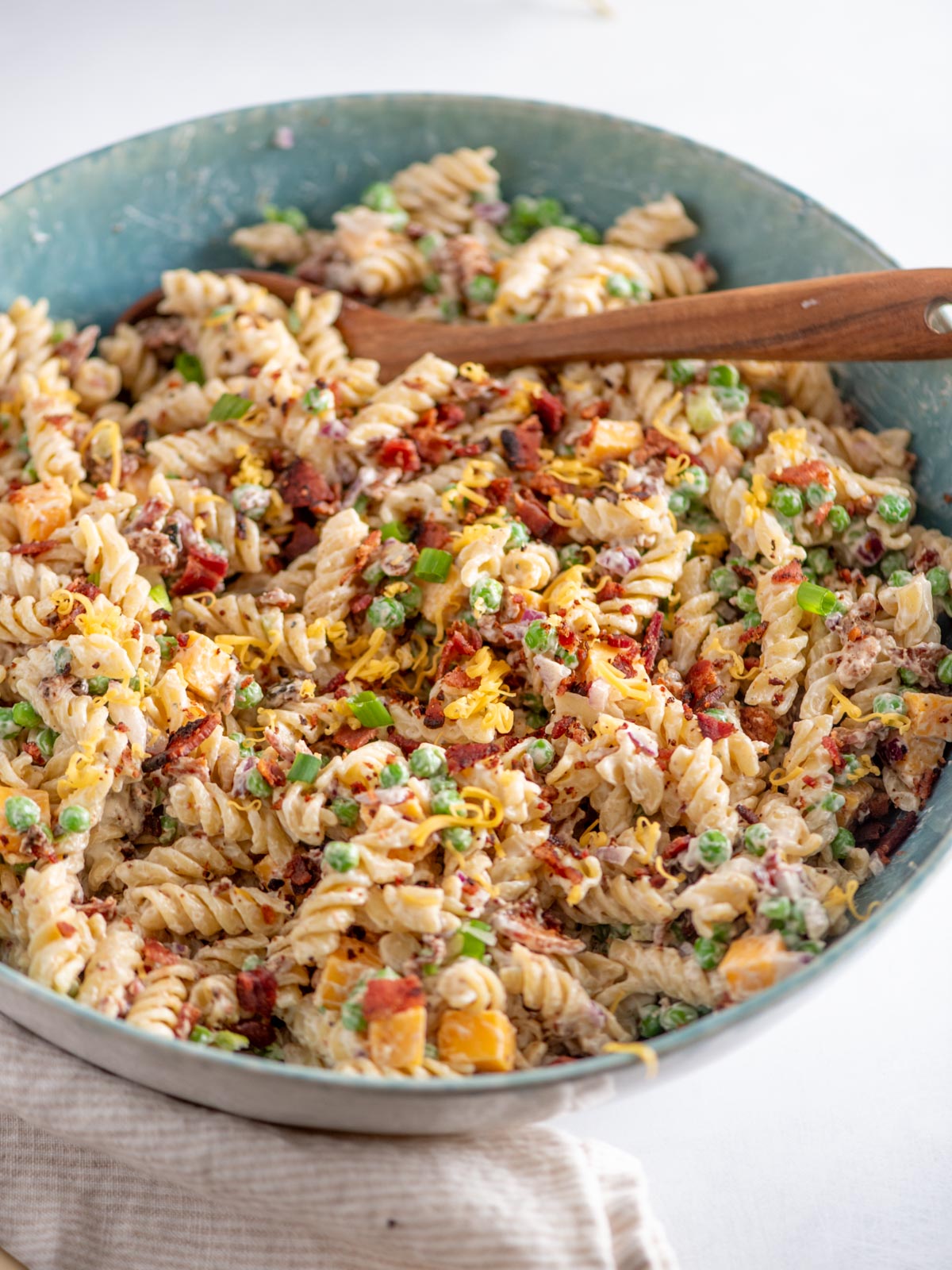 A large bowl of creamy rotini pasta mixed with peas, sun-dried tomatoes, chopped vegetables, and herbs, topped with shredded cheese, with a wooden serving spoon resting in the bowl.