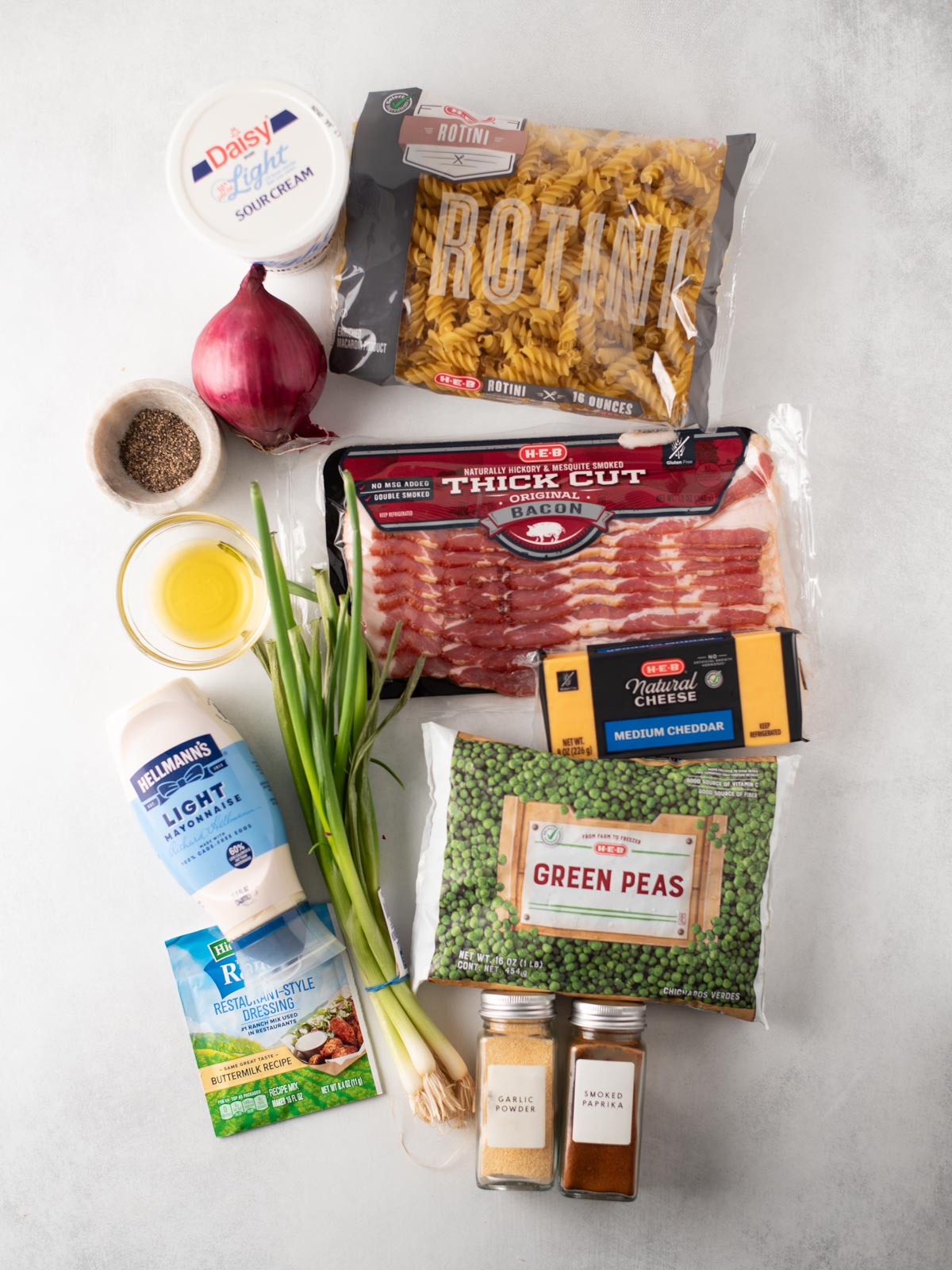 A flat lay of ingredients on a white surface: rotini pasta, sour cream, red onion, black pepper, olive oil, green onions, bacon, cheddar cheese, green peas, mayonnaise, ranch seasoning, garlic, and two spice jars.