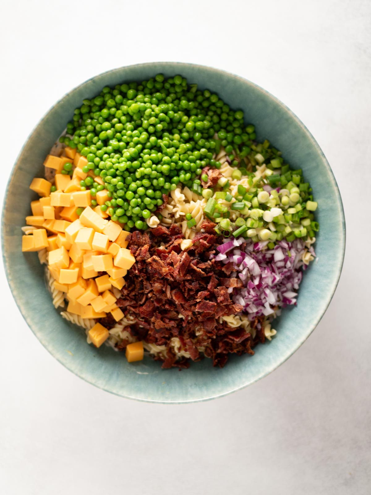 A blue bowl filled with separated ingredients: green peas, chopped green onions, cubed cheddar cheese, chopped red onion, crispy bacon bits, and cooked pasta spirals on a light background.