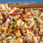 A close-up of bacon ranch pasta salad with rotini pasta, bacon bits, shredded cheese, chopped green onions, peas, and ranch dressing, topped with more bacon and cheese. Text above reads Bacon Ranch Pasta Salad.