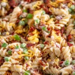 Close-up of a bowl filled with creamy rotini pasta salad mixed with bacon, cheddar cheese, peas, and green onions. A banner at the top reads bacon ranch pasta salad with sweetlysplendid.com below.