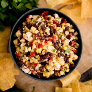 A bowl of vibrant black bean corn feta dip—with red pepper and chopped herbs—sits surrounded by crisp tortilla chips on a rustic wooden surface.