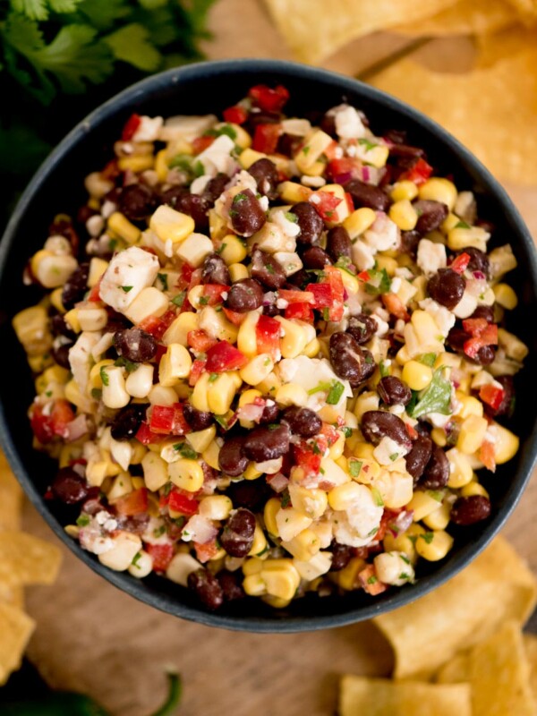A bowl of vibrant black bean corn feta dip—with red pepper and chopped herbs—sits surrounded by crisp tortilla chips on a rustic wooden surface.