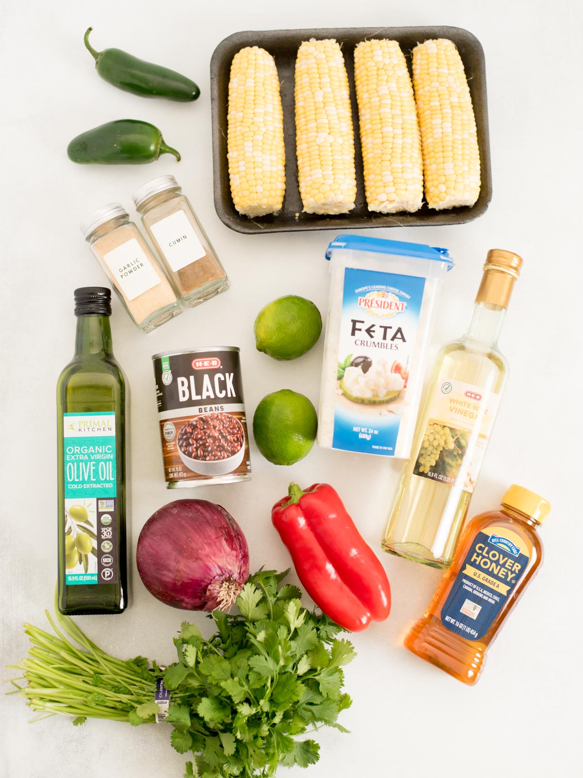 Ingredients for black bean corn feta dip arranged on a white surface: corn, jalapeños, spices, feta, black beans, limes, olive oil, vinegar, honey, red onion, red bell pepper, and fresh cilantro.