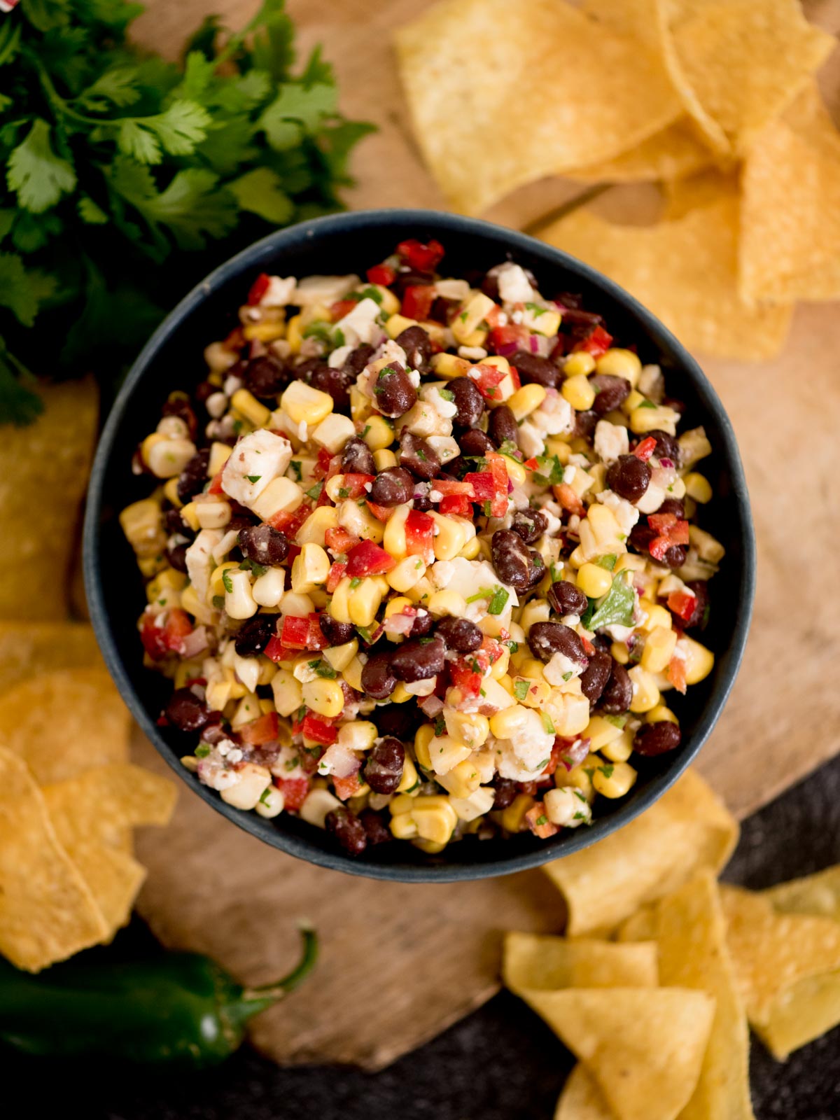 A bowl of colorful black bean corn feta dip with diced red peppers, surrounded by tortilla chips on a wooden surface.
