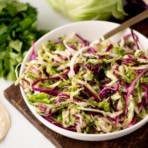 A bowl of fresh coleslaw with shredded green and purple cabbage and herbs, sitting on a wooden board with a bunch of cilantro and a cabbage in the background.