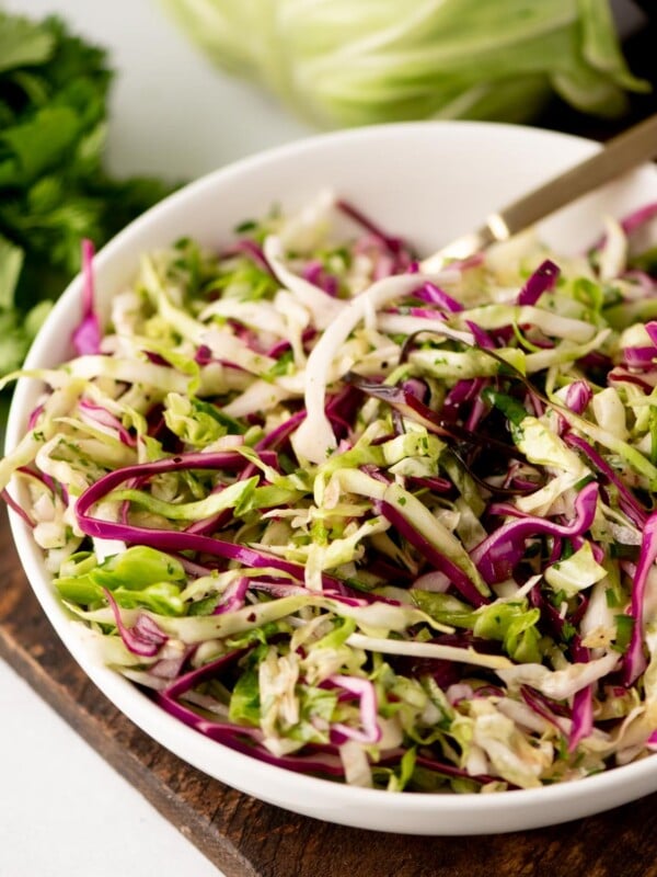 A bowl of fresh coleslaw with shredded green and purple cabbage and herbs, sitting on a wooden board with a bunch of cilantro and a cabbage in the background.