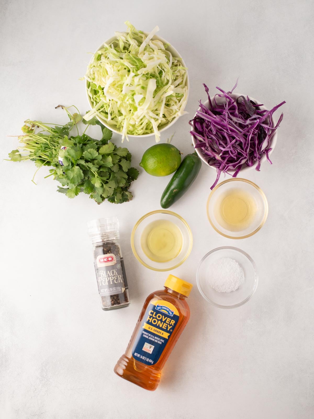 Overhead view of ingredients on a white surface: shredded green and purple cabbage, cilantro, a lime, jalapeño, black pepper, honey, salt, white vinegar, and oil in small bowls.