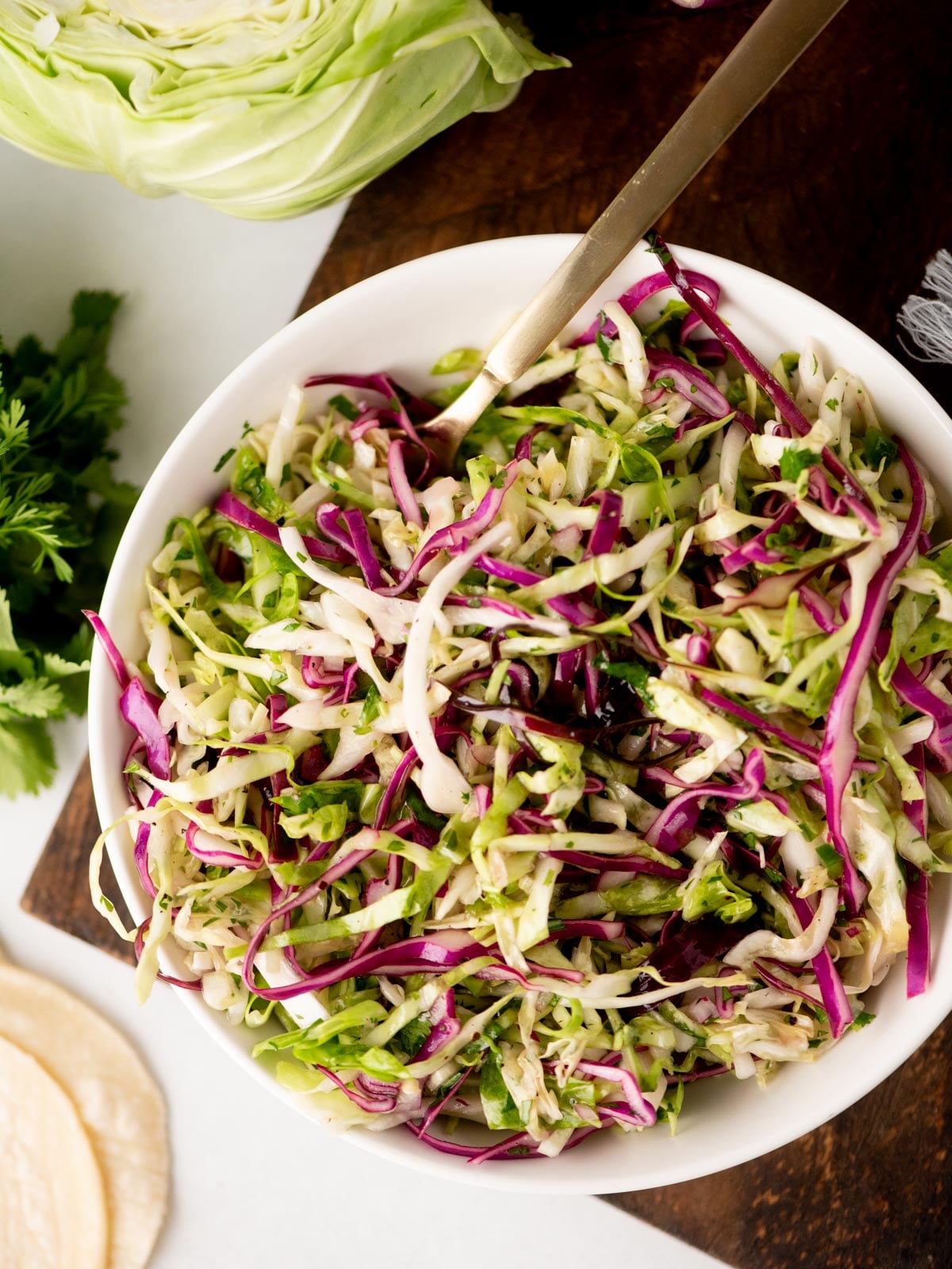 A bowl of shredded cabbage slaw with green and purple cabbage and herbs, served on a wooden surface with a gold fork. Fresh cabbage, parsley, and tortillas are visible nearby.