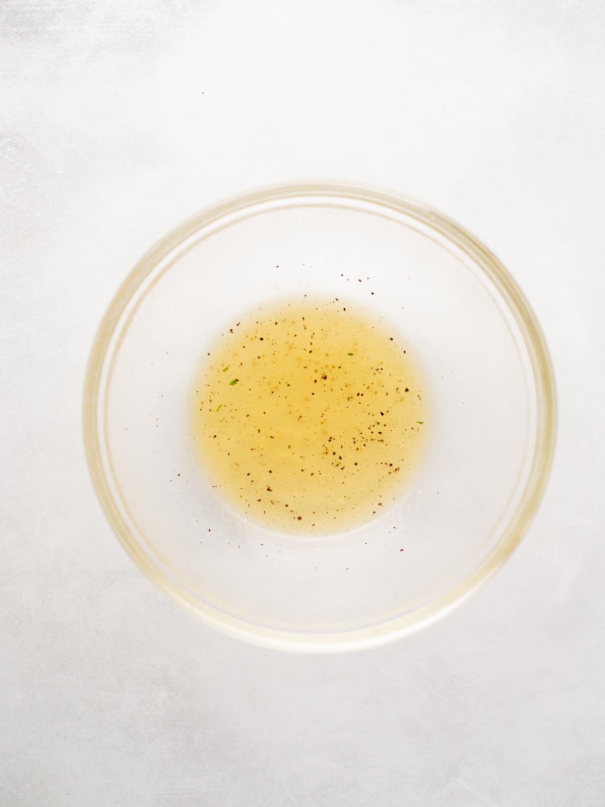 A clear glass bowl containing a small amount of light yellow liquid with specks of black pepper, placed on a white surface.