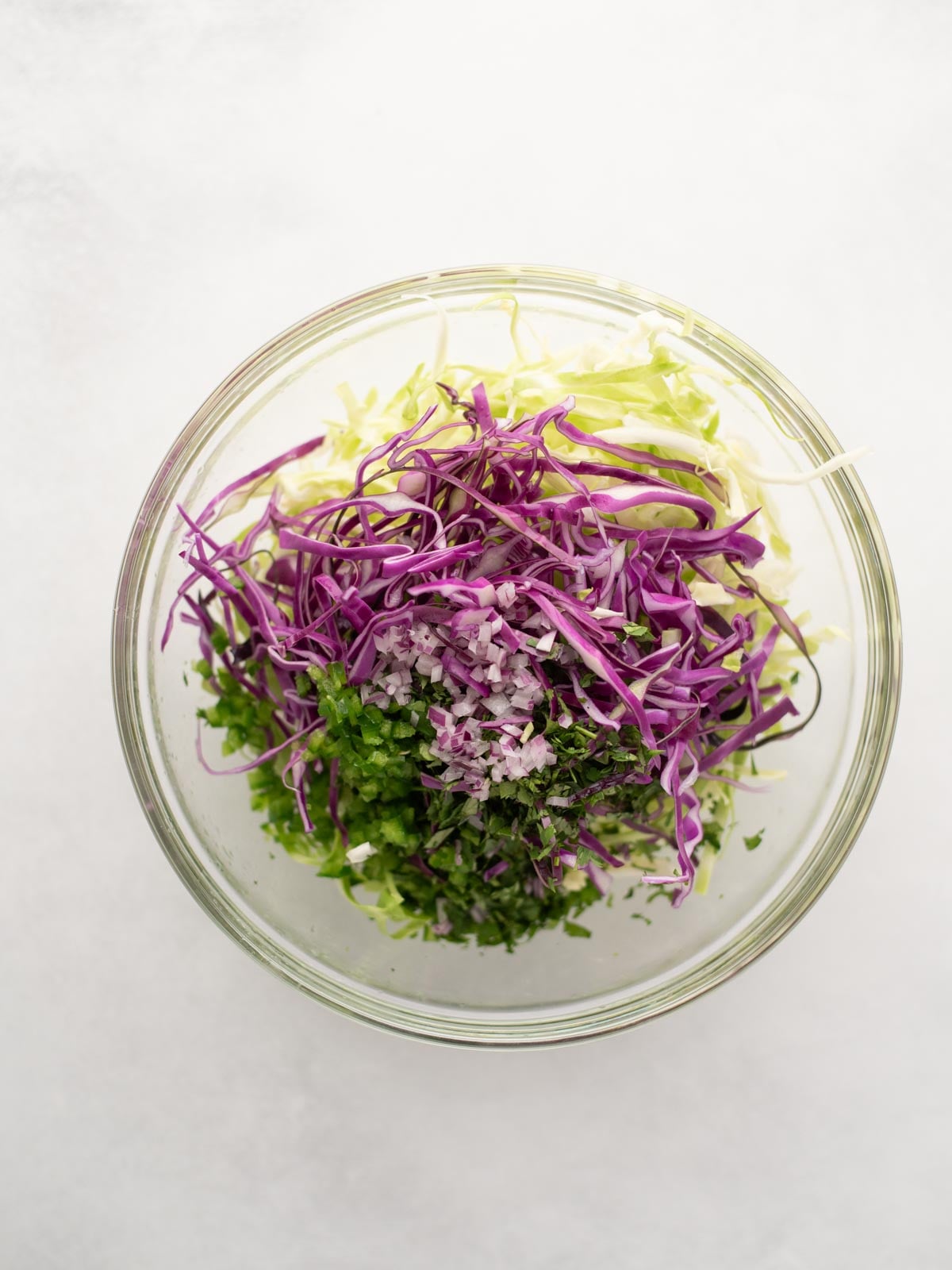 A glass bowl filled with shredded green cabbage, shredded purple cabbage, chopped cilantro, and finely diced red onion, all arranged in separate sections on a white background.