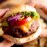 A close-up of hands holding a cheeseburger with a beef patty, melted cheddar cheese, tomato slices, red onion, pickles, and lettuce in a bun.