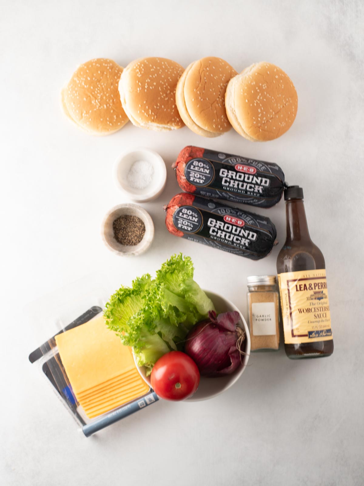 Top-down view of burger ingredients: four sesame seed buns, two rolls of ground chuck, salt, pepper, Worcestershire sauce, garlic powder, cheese slices, lettuce, a tomato, and a red onion on a white surface.