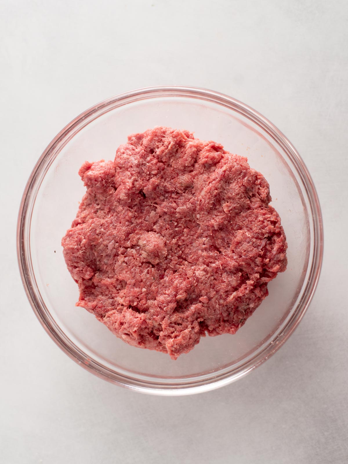A clear glass bowl filled with raw ground beef sits on a white surface.