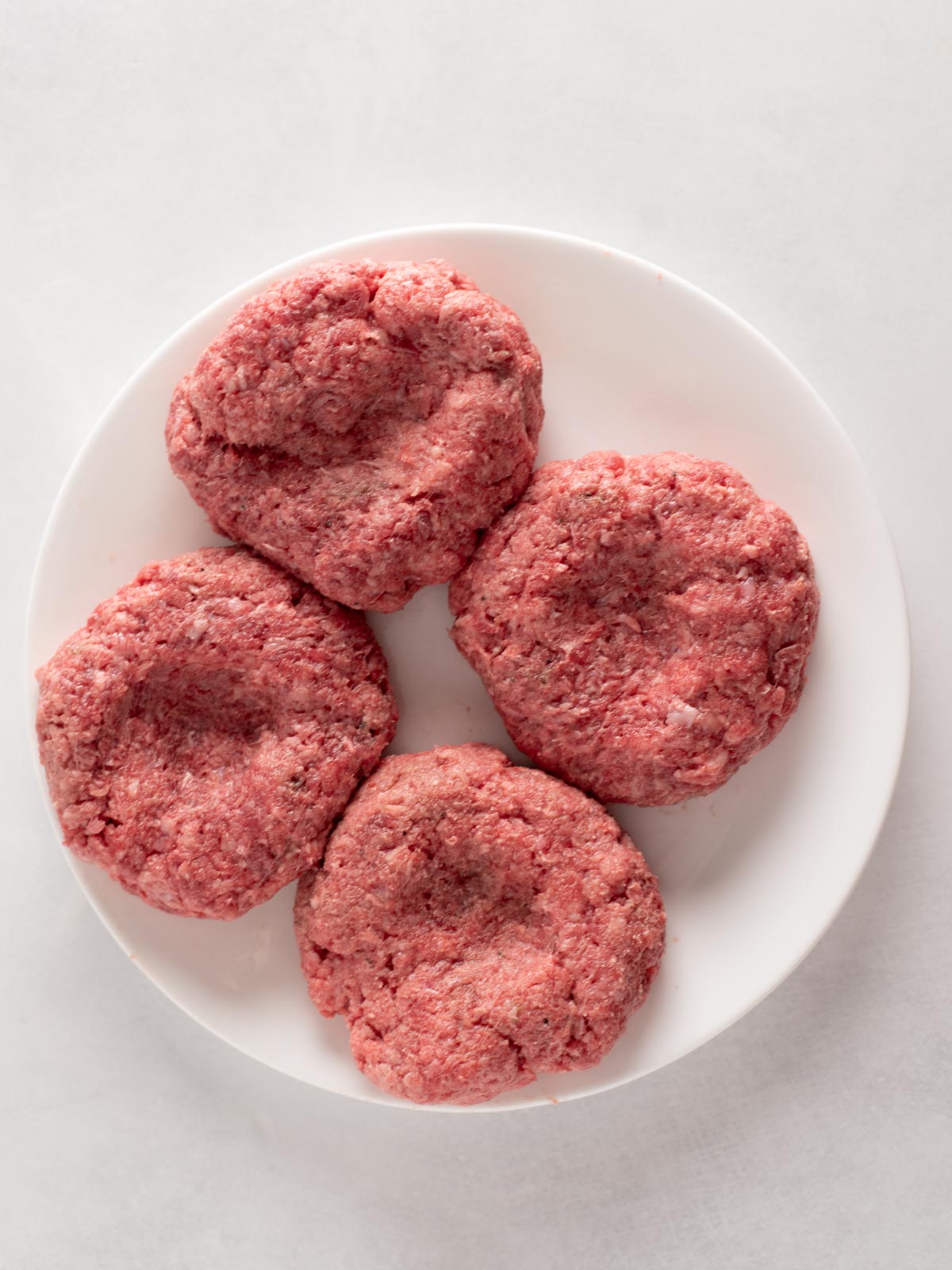 Four raw ground beef patties arranged in a circle on a white plate, with visible indentations in the center of each patty, set against a light background.