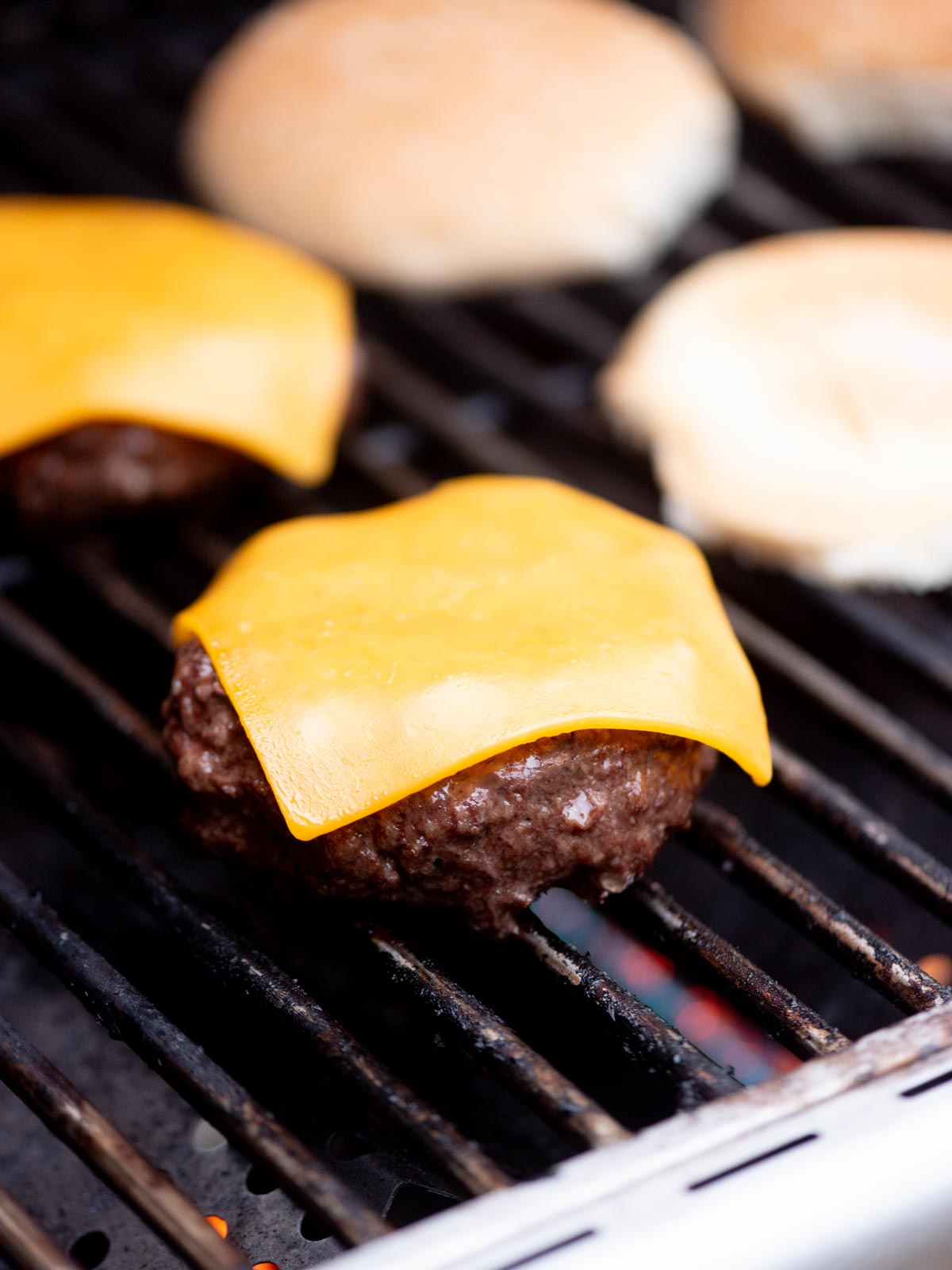 A close-up of a beef burger patty with melted cheddar cheese on a grill, with two burger buns toasting in the background.