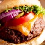 A close-up of a grilled beef burger topped with melted cheddar cheese, lettuce, tomato, pickles, and red onion in a sesame bun. Text above reads: the best grilled burgers, sweetlysplendid.com.