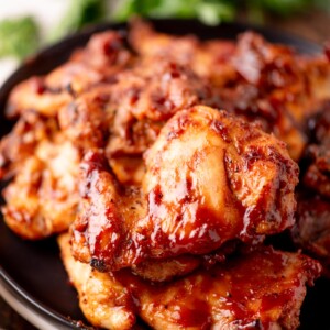 Close-up of a plate filled with glazed, grilled chicken pieces, coated in a rich, shiny barbecue sauce, with a blurred green garnish in the background.