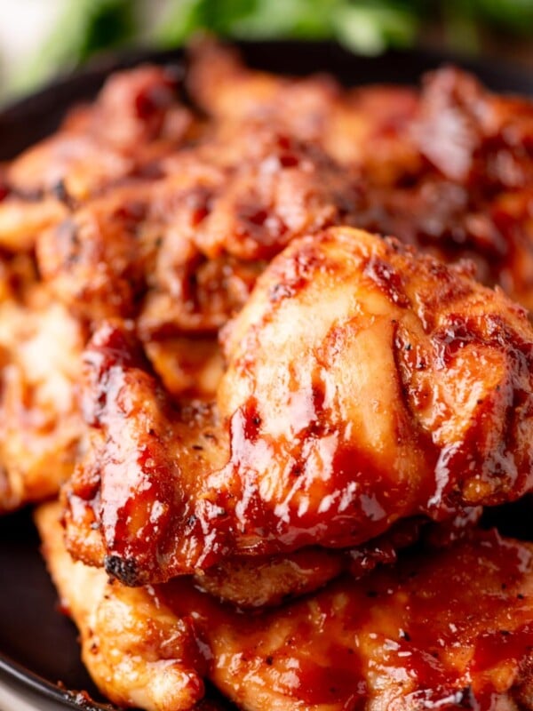 Close-up of a plate filled with glazed, grilled chicken pieces, coated in a rich, shiny barbecue sauce, with a blurred green garnish in the background.